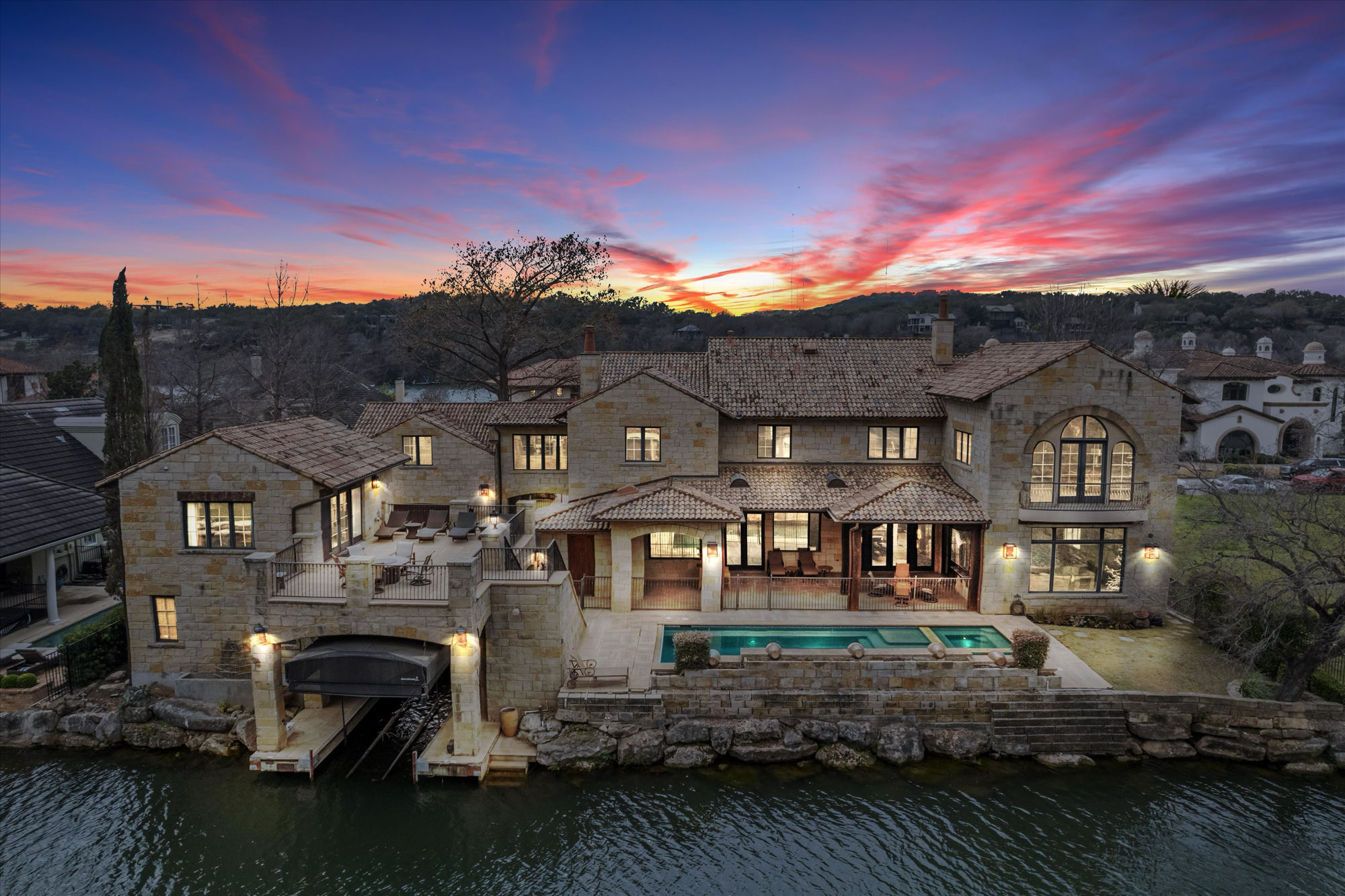 Twilight aerial view of the lakefront stone mansion with infinity pool, spa, and covered boat dock on Lake Austin