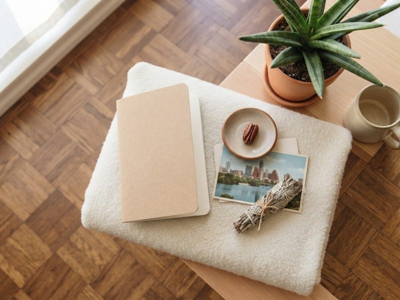 A notebook, a postcard showing a city skyline, a sage bundle, a small ceramic dish with a pecan, a potted succulent, and a mug on a table next to parquet flooring.