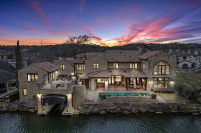 Twilight aerial view of the lakefront stone mansion with infinity pool, spa, and covered boat dock on Lake Austin