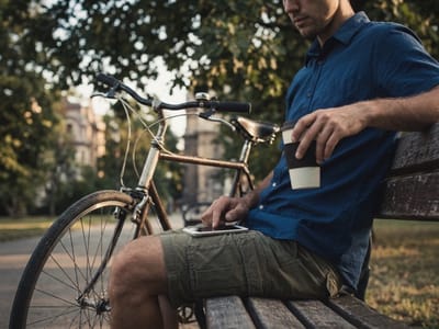 Man with a coffee cup and phone sitting on a park bench beside his bicycle