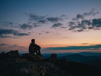 Person sitting alone on a rocky mountaintop watching a dusky sunset over distant ridges