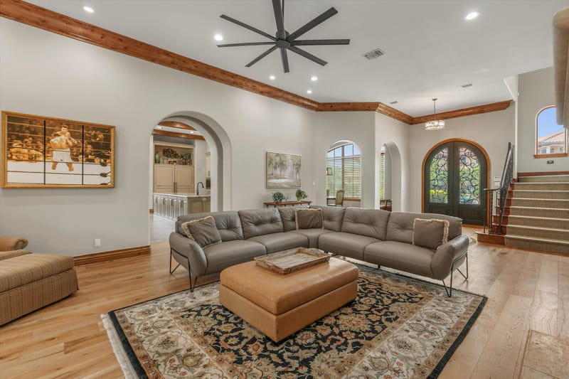 Open-concept living room with leather sectional, framed Muhammad Ali artwork, wood-beam ceiling, and arched iron-and-glass front doors