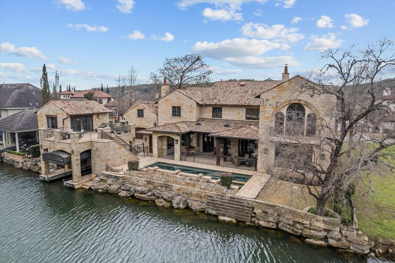 Daytime aerial of the stone waterfront home with pool, terraces, and a covered boat dock